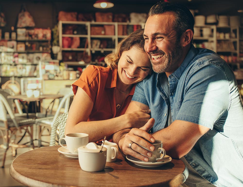 This is a photo of a happy couple laughing in a cafe. The woman is leaning over to hug the man.